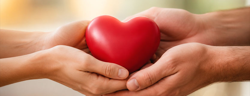 Mani di una coppia che custodiscono insieme un cuore rosso, simbolo dell’amore, dell’intimità e della complicità.