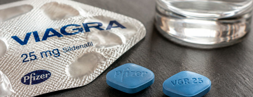 Compresse blu di Viagra accanto alla confezione del farmaco e a un bicchiere d’acqua.