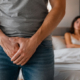 Uomo vestito con jeans e maglietta tiene le mani davanti all’inguine mentre la partner è seduta sul letto sullo sfondo.