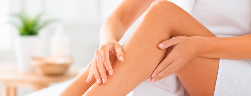 Donna adulta avvolta in un asciugamano accarezza delicatamente la pelle della propria gamba in un ambiente luminoso dedicato alla cura del corpo.