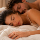 Coppia adulta distesa a letto, con la donna in primo piano e l’uomo alle sue spalle, entrambi con gli occhi chiusi.