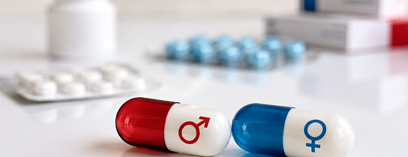 Due capsule farmaceutiche con simboli maschile e femminile su un piano bianco, con confezioni di medicinali sullo sfondo.