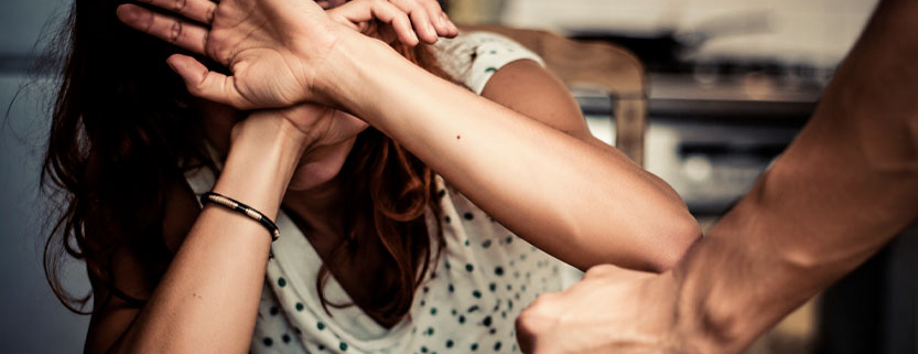 Donna adulta impaurita si protegge il volto con le braccia mentre un uomo la minaccia con un pugno.