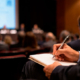 Medico prende appunti durante un congresso scientifico con relatori e pubblico in sala.