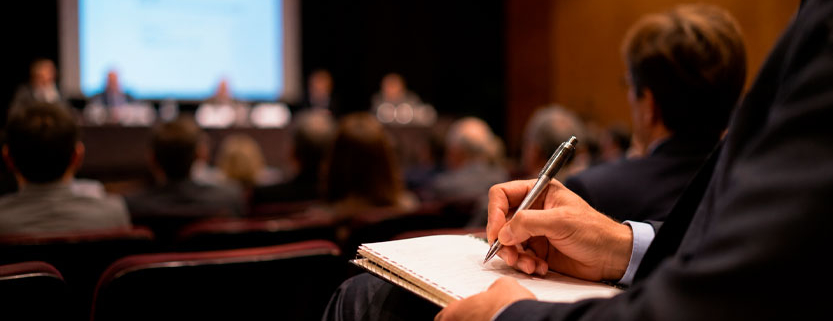 Medico prende appunti durante un congresso scientifico con relatori e pubblico in sala.