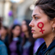 Donna adulta di profilo con il simbolo femminile dipinto sulla guancia, tra altre persone sullo sfondo.