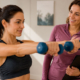 Donna adulta si allena con i manubri in palestra sotto la supervisione di una personal trainer.
