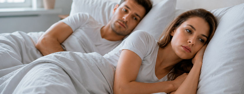 Giovane coppia adulta a letto con la donna assorta e distante mentre l'uomo la osserva con preoccupazione.