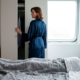 Un uomo a letto a torso nudo guarda verso il soffitto con le mani dietro la testa, mentre una donna in vestaglia di seta blu guarda dentro l'armadio aperto.