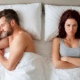 Vista dall'alto di un uomo a letto di profilo ad occhi chiusi e di una donna a fianco a braccia conserte con espressione imbronciata