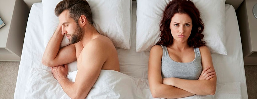 Vista dall'alto di un uomo a letto di profilo ad occhi chiusi e di una donna a fianco a braccia conserte con espressione imbronciata