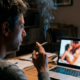 Un uomo di circa 38 anni seduto alla scrivania di notte, illuminato dallo schermo del computer con una sigaretta accesa in mano.