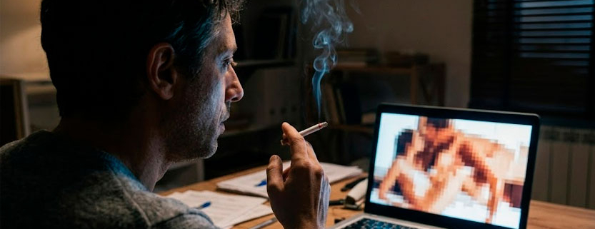 Un uomo di circa 38 anni seduto alla scrivania di notte, illuminato dallo schermo del computer con una sigaretta accesa in mano.