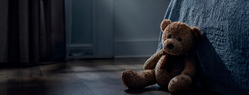 Orsetto di peluche abbandonato sul pavimento accanto a un letto in una stanza buia