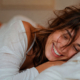 Donna adulta spettinata e sorridente distesa sul letto con il partner sfocato sullo sfondo