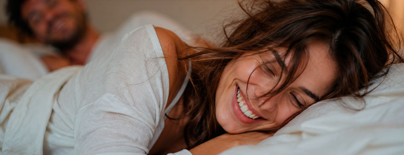 Donna adulta spettinata e sorridente distesa sul letto con il partner sfocato sullo sfondo