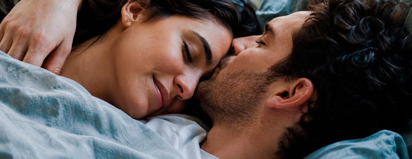 Coppia adulta abbracciata a letto mentre l’uomo bacia teneramente la donna sul viso